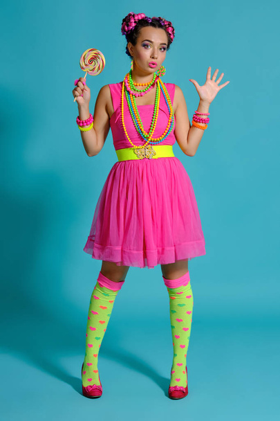 Lovely girl with a multi-colored braids hairstyle and bright make-up, posing in studio against a blue background, holding a lollipop in her hand. - Foto, imagen