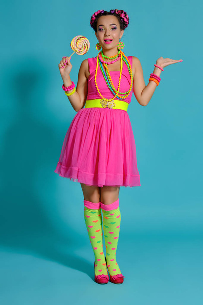 Lovely girl with a multi-colored braids hairstyle and bright make-up, posing in studio against a blue background, holding a lollipop in her hand. - Valokuva, kuva