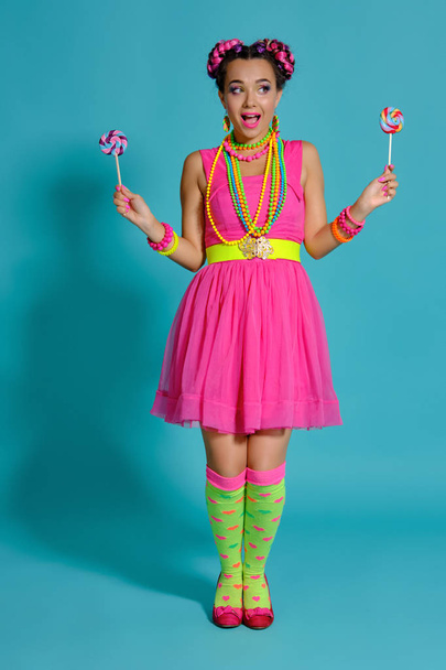Lovely girl with a multi-colored braids hairstyle and bright make-up, posing in studio against a blue background, holding a lollipop in her hand. - Photo, Image