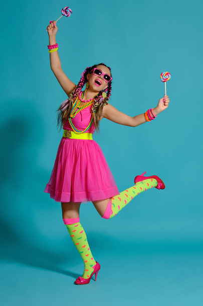 Lovely girl with a multi-colored braids hairstyle and bright make-up, posing in studio against a blue background, holding a lollipop in her hand. - Foto, Imagem