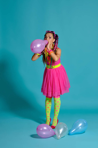 Krásná dívka s pestrobarevnými copánky účes a světlý make-up, pózování ve studiu s vzduchovými balónky na modrém pozadí. - Fotografie, Obrázek