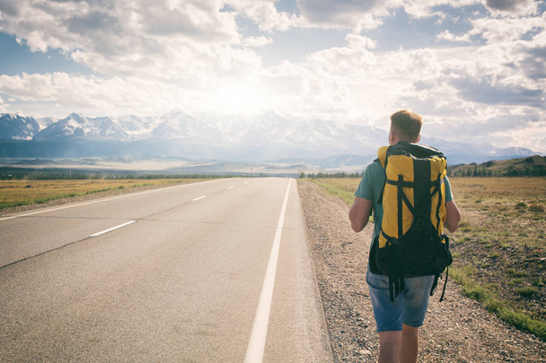 Adam sırtında dağlara bakan bir sırt çantası ile bir asfalt yol boyunca yürüyor - Fotoğraf, Görsel