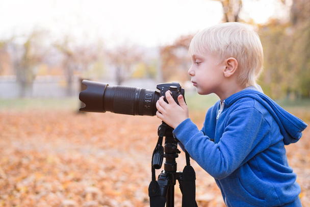 Πορτραίτο ενός μικρού ξανθού αγοριού με μια μεγάλη κάμερα DSLR σε τρίποδο. Φωτογραφία συνεδρία στο φθινοπωρινό πάρκο - Φωτογραφία, εικόνα