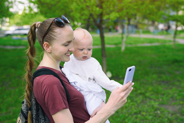 Młoda matka z dzieckiem w ramionach i używa smartfona. Selfie z dzieckiem. Park sprężynowy - Zdjęcie, obraz