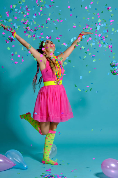 Menina encantadora com um penteado de tranças multi-coloridas e maquiagem brilhante, posando em estúdio com balões de ar e confete contra um fundo azul
. - Foto, Imagem