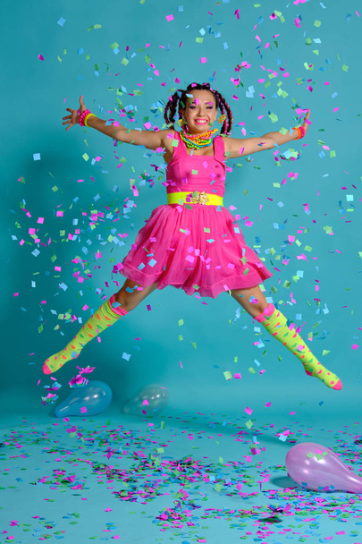 Lovely girl with a multi-colored braids hairstyle and bright make-up, posing in studio with air balloons and confetti against a blue background. - Foto, Imagem