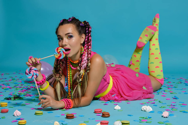 Lovely girl with a multi-colored braids hairstyle and bright make-up, posing in studio with lollipop, air balloons and confetti against a blue background. - Fotoğraf, Görsel