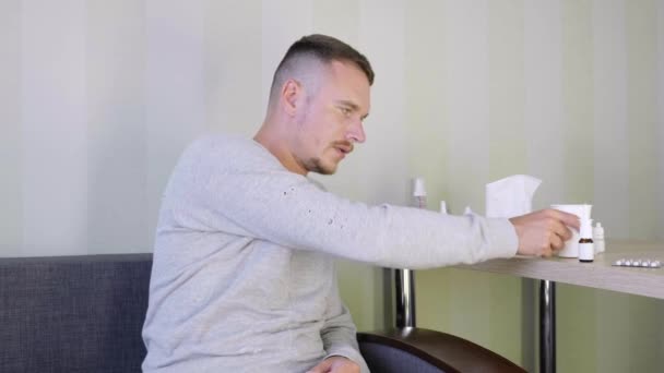 Sick man is self-medicating at home. Upset young guy drinking drugs from a paper cup. Plenty pills and medications are on the bedside table. Acute Respiratory Virus Infection. Illness cold flu. 4K - Footage, Video