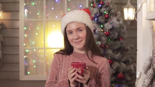 Pretty woman in red hat with cup of tea smiling and looking at camera. Christmas concept. Close-up view - Materiał filmowy, wideo