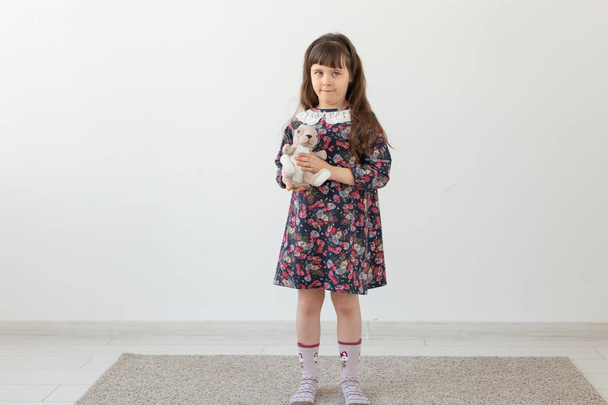 Маленька чарівна дівчинка в квітковій сукні м'яко обіймає свого улюбленого іграшкового кролика, що стоїть на білому тлі. Концепція дитячих іграшок та товарів
 - Фото, зображення