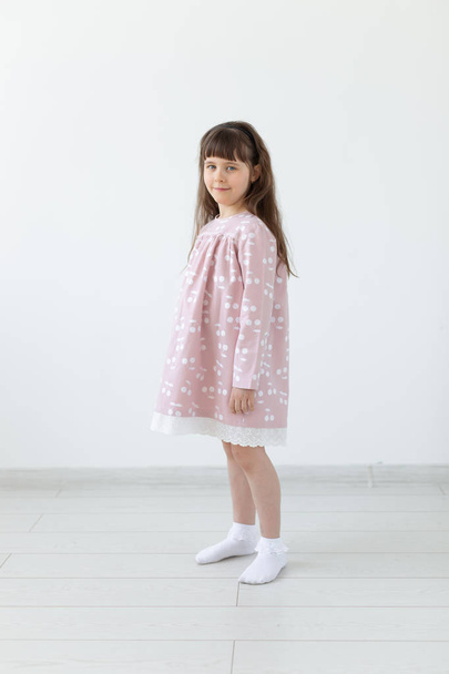 Kleine trotse brunette meisje in een roze jurk staat op een witte achtergrond. Het concept van stijlvolle kinderkleding en voorschoolse leeftijd. Copyspace. - Foto, afbeelding