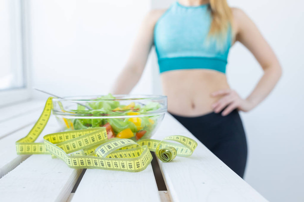 gesunder Lebensstil, Fitness und Ernährungskonzept - Diätsalat aus nächster Nähe und Maßband mit Frau in Sportkleidung im Hintergrund - Foto, Bild