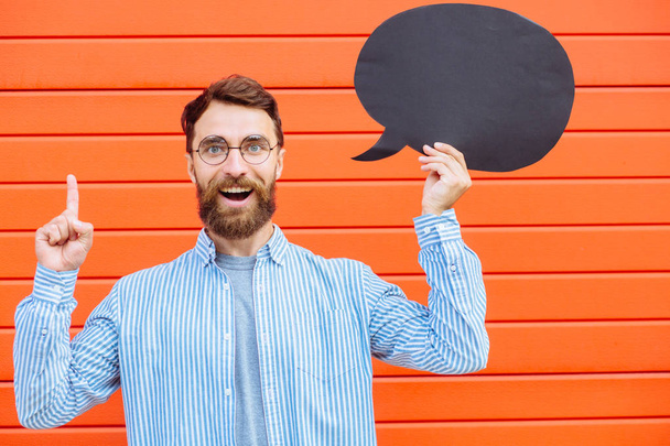 Attractive young beard man in circle glasses with fun fantasy emotion on his face holding a yellow speech bubble looking at camera and smiling, on red background outdoor portrait. - Foto, Bild