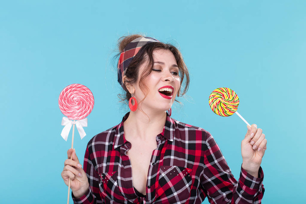 Charming young woman in retro clothes holding colorful lollipops in her hands and licking one posing on blue background. Concept of love for sweets and holidays. - 写真・画像