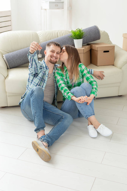 Charming young woman kisses her husband holding in hands the keys to their new apartment while sitting in her new living room. Housewarming and mortgage in new building concept. - Foto, imagen