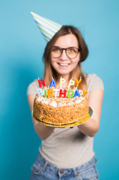 Close-up grappig positief meisje in glazen en groet papier hoed houden een gelukkige verjaardagstaart in haar handen staande op een blauwe achtergrond. - Foto, afbeelding
