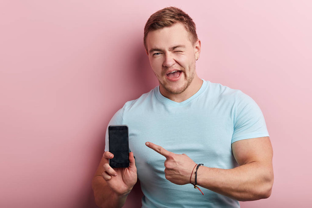 happy strong muscular smiling man in stylish blue T-shirt showing smartphone - Fotoğraf, Görsel