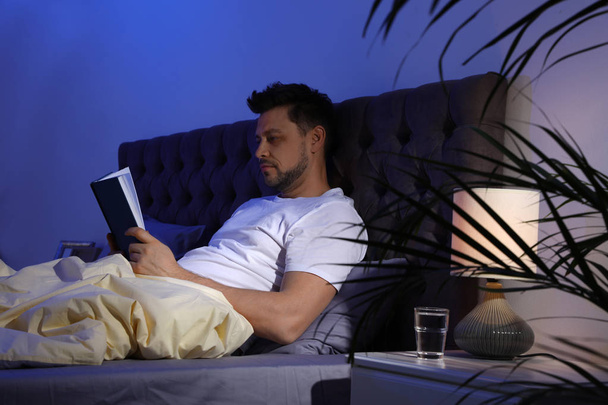 Handsome man reading book in dark room at night. Bedtime - Φωτογραφία, εικόνα