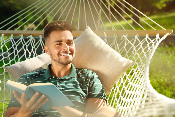 Young man reading book in comfortable hammock at green garden - Foto, Imagen