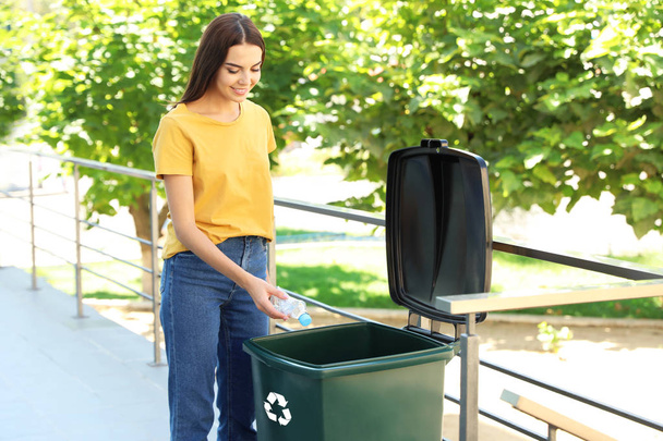 Young woman throwing plastic bottle into recycling bin outdoors - 写真・画像