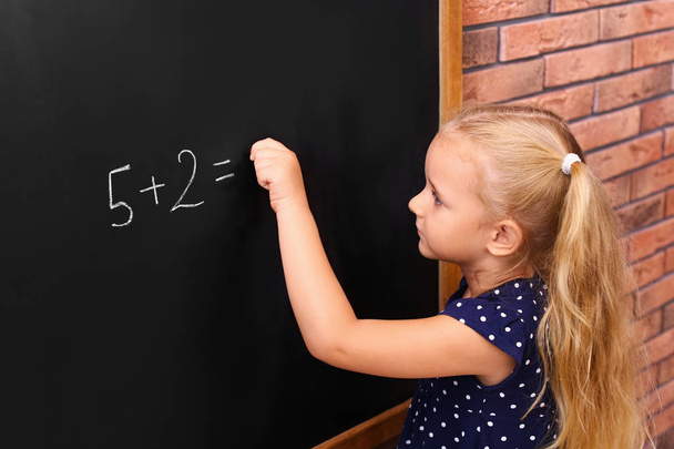 Cute little left-handed girl doing sums on chalkboard near brick wall - Photo, Image