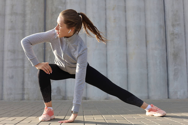 Young attractive sporty girl warming up outdoors, doing stretching at sunset or sunrise in city - Photo, Image