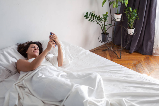 Frau mit Smartphone im Bett mit weißen Laken - Foto, Bild