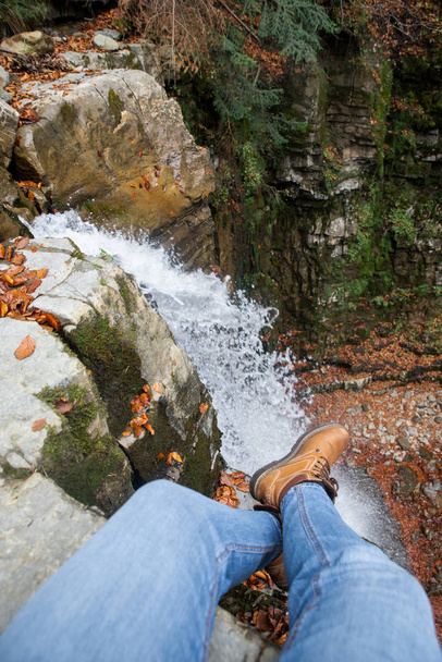 piernas de hombre en pantalones vaqueros azules y botas marrones sentados en el borde mirando la cascada
 - Foto, Imagen