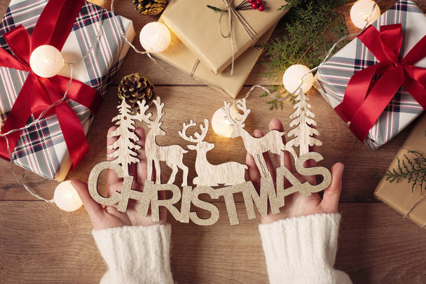 Female Hands Holding Merry Christmas Inscription, Fir Tree, Figures of Deers And Festive Gift Boxes. Winter Holidays And Happy New Year Concept. Flat Lay Of Christmas Accessories And Lights - Foto, imagen