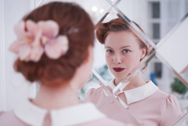 Κορίτσι σε vintage φόρεμα κοιτάζοντας στον καθρέφτη χαρούμενο και χαρούμενο. Χαριτωμένο νεαρή γυναίκα σε ροζ ρετρό φόρεμα μόνο στο λευκό δωμάτιο. - Φωτογραφία, εικόνα
