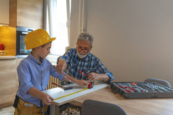 Grandfather and grandson in workshop. Grandfather and grandson crafting wooden materials. My grandson is little carpenter. Grandfather Teaching Boy Carpentry  - 写真・画像