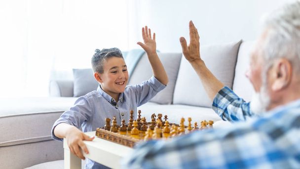 Happy boy grandson playing chess with old senior man grandfather at home. Side view of happy grandson and grandfather looking at each other while playing chess at home - Foto, imagen