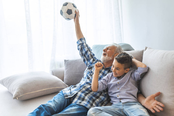 Excited grandchild and senior man watching football match. Overjoyed grandfather and kid watching football match together, cheering for a football game and holding a football ball  - Zdjęcie, obraz