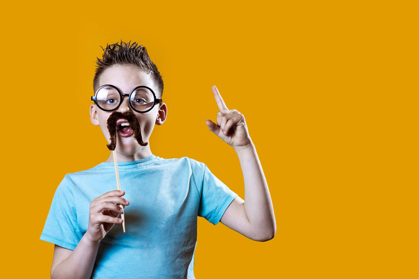 een jongen in een licht t-shirt met een snor en bril houdt zijn wijsvinger omhoog tegen felle achtergrond - Foto, afbeelding