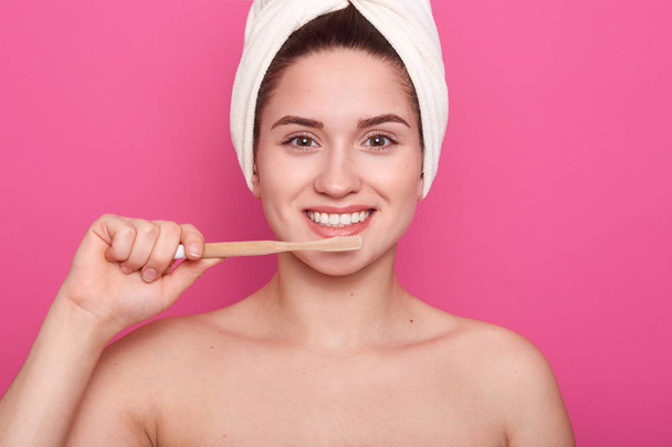 Gros plan portrait de jolie femme souriante caucasienne isolée sur studio rose, se brosser les dents dans la salle de bain, faire la procédure du matin avec une serviette blanche sur la tête, exprime la joie. Concept de santé
 - Photo, image
