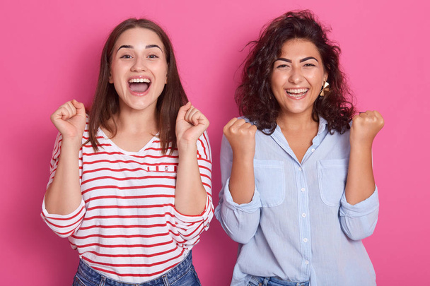 Two beautiful girlfriend girls happily and enthusiastically squeeze their fists to worry about their favorite team. The joy of victory and happiness. Studio portrait on pink isolated background - Foto, imagen
