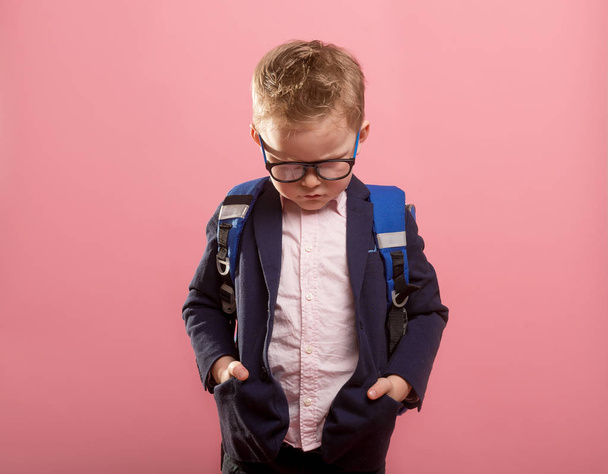 Шкільний хлопчина нервово дивиться вниз. Дитячий хлопчик у формі, окуляри та рюкзак ізольовані на рожевому. Сан і нещасний портрет Кіда. Знову в школу - Фото, зображення