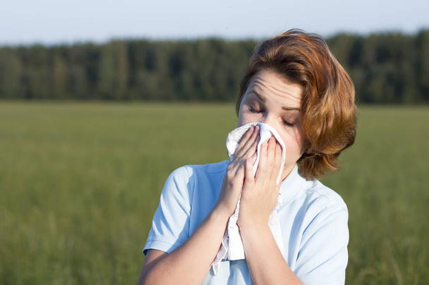 Young woman blowing her nose with tissue, snot in a paper handkerchief outdoor. Sick girl sneezes with eyes closed in a summer field. Natural background. Feeling unwell, ill, bad, suffering. Allergy.  - Foto, immagini
