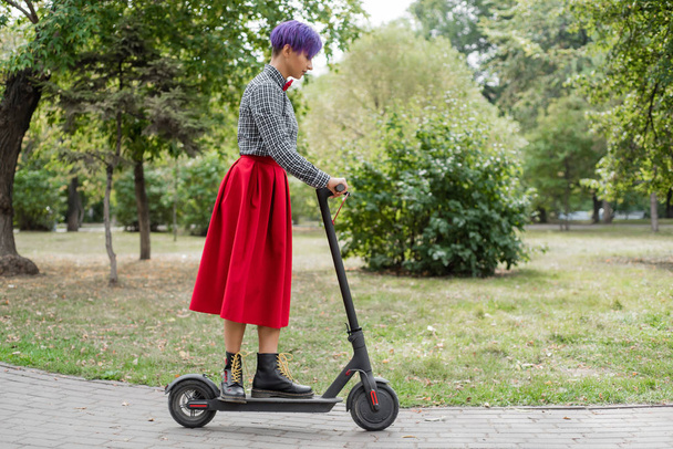 A young woman with purple hair rides an electric scooter in a park. A stylish girl with a shaved temple in a plaid shirt, a long red skirt and a bow tie is riding around the city on a modern device.  - 写真・画像