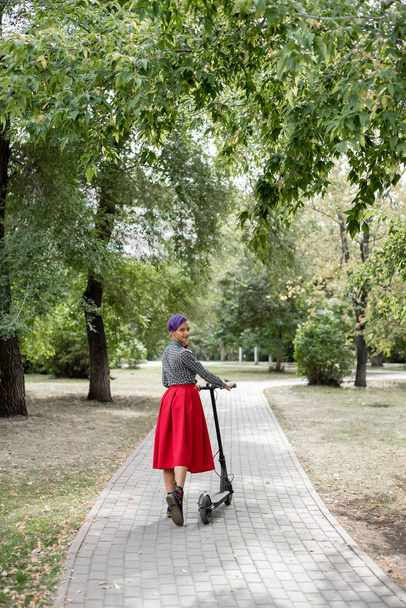 A young woman with purple hair rides an electric scooter in a park. Stylish girl in a plaid shirt, a long red skirt and a bow tie is riding around the city on a modern device. Hipster. - Foto, Imagen