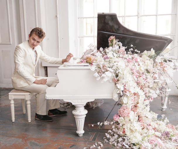 花の大きな白いピアノを持つ白いビジネススーツを着た若い男のティーンエイジャー。白人男性は休日だけで音楽を演奏. - 写真・画像