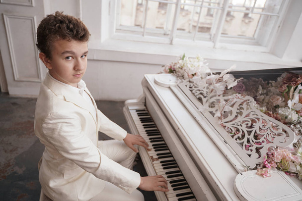 νέος άνδρας Έφηβος με μια λευκή επαγγελματική στολή με ένα μεγάλο λευκό πιάνο στα λουλούδια. Καυκάσιος αρσενικό παίζουν μουσική μόνο στις διακοπές. - Φωτογραφία, εικόνα