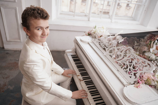 młody mężczyzna nastolatek w białym garniturze biznesowym z dużym białym fortepianem w kwiaty. kaukaski mężczyzna grać muzykę na wakacjach sam. - Zdjęcie, obraz