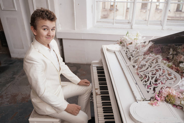 jonge man tiener in een wit Business Suit met een grote witte piano in bloemen. blanke man spelen muziek bij vakantie alleen. - Foto, afbeelding