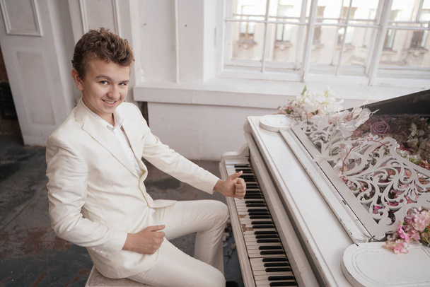 joven adolescente en un traje de negocios blanco con un gran piano blanco en flores. caucásico macho jugar música en vacaciones solo
. - Foto, imagen