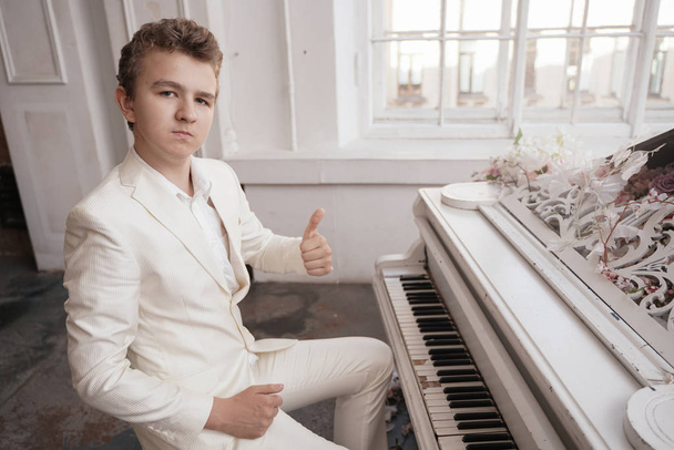 young man teenager in a white business suit with a large white piano in flowers. caucasian male play music at holiday alone. - Фото, изображение