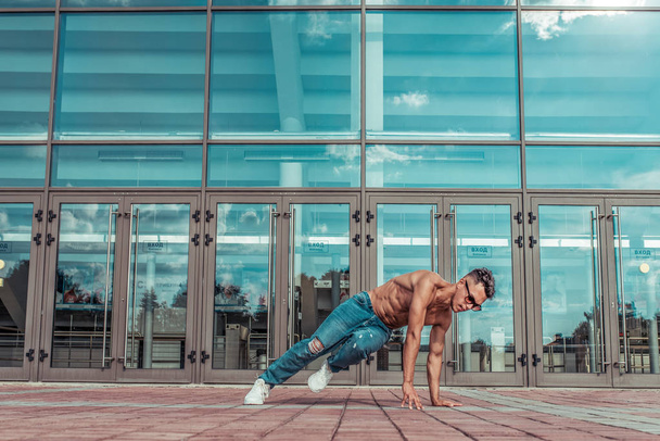 Young stylish athlete background glass windows, guy dancer, summer city dancing break dance, healthy fitness athlete life style, jeans sneakers. Dancing music. Performs public. Free space text. - Photo, Image