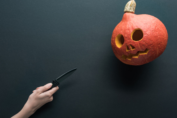 cropped view of woman holding knife in front of spooky Halloween pumpkin on black background - Photo, Image