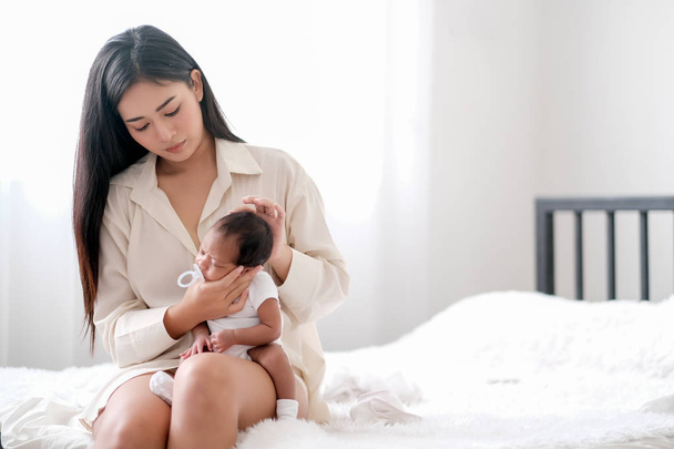 weiße Hemd asiatische Mutter kümmern sich um ihr Neugeborenes und sitzen auf dem Bett vor Vorhang mit Tageslicht und Kopierraum. Sie zeigt auch ihre Liebe zum Baby. - Foto, Bild