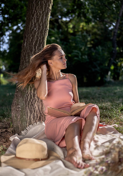 Beautiful girl resting on the grass by the tree. In a hat with a book of dried flowers and a smartphone. - Foto, Imagen